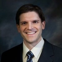 Professional headshot of a man with dark hair smiling at the camera and wearing a suit and tie.